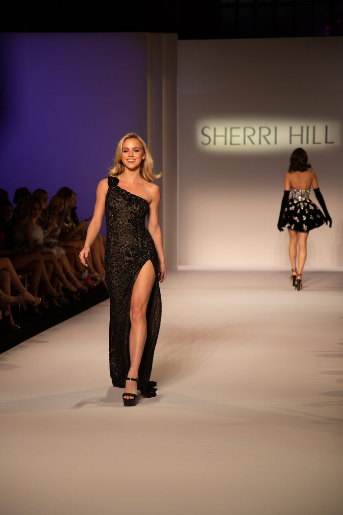 Model Emma Brooks in the black one-shoulder gown with a high slit on the runway, with the silver beaded mini behind