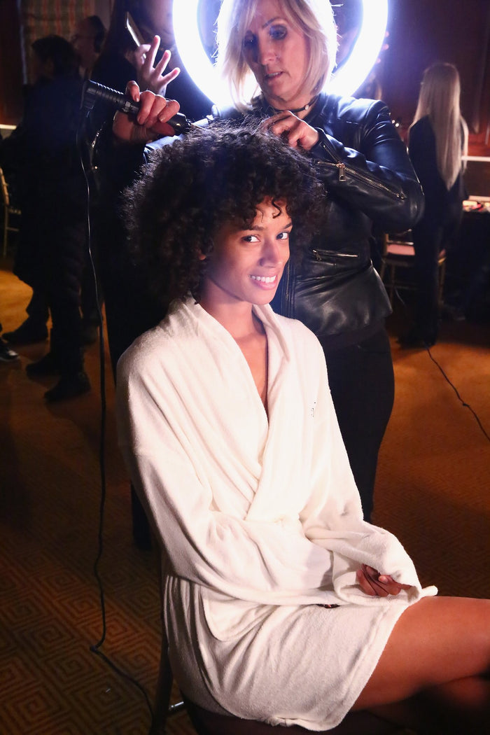 A model in a white robe has her hair finished backstage