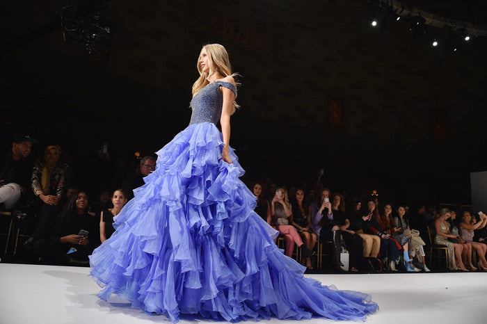 A periwinkle ruffled ball gown comes down the runway, seen from the front row