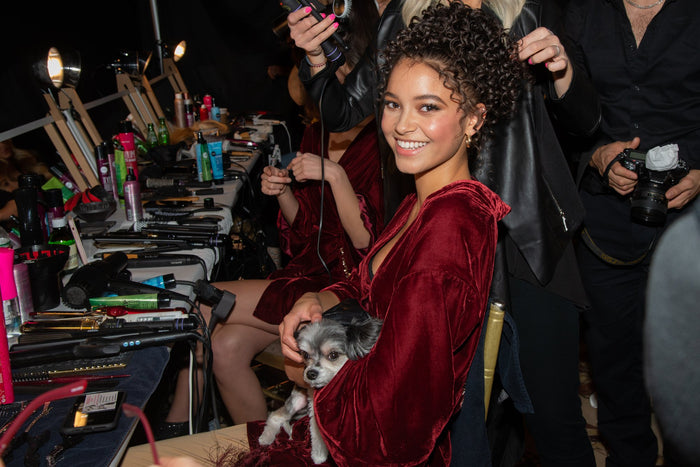 A model in a burgundy velvet robe at the makeup table with a small dog on her lap