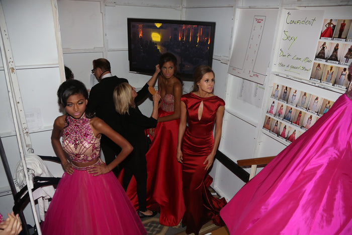 Models in hot pink and red gowns lining up backstage beside the look board