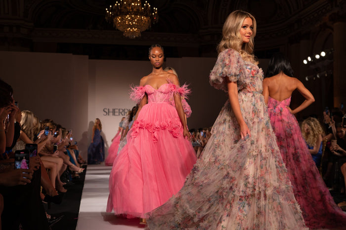 Pink and floral gowns pass on the runway, seen from the front row